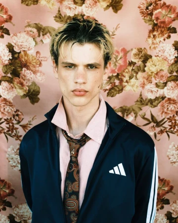 Model Lazar Dedic in a track jacket, pink shirt, and paisley tie against a pink floral background.