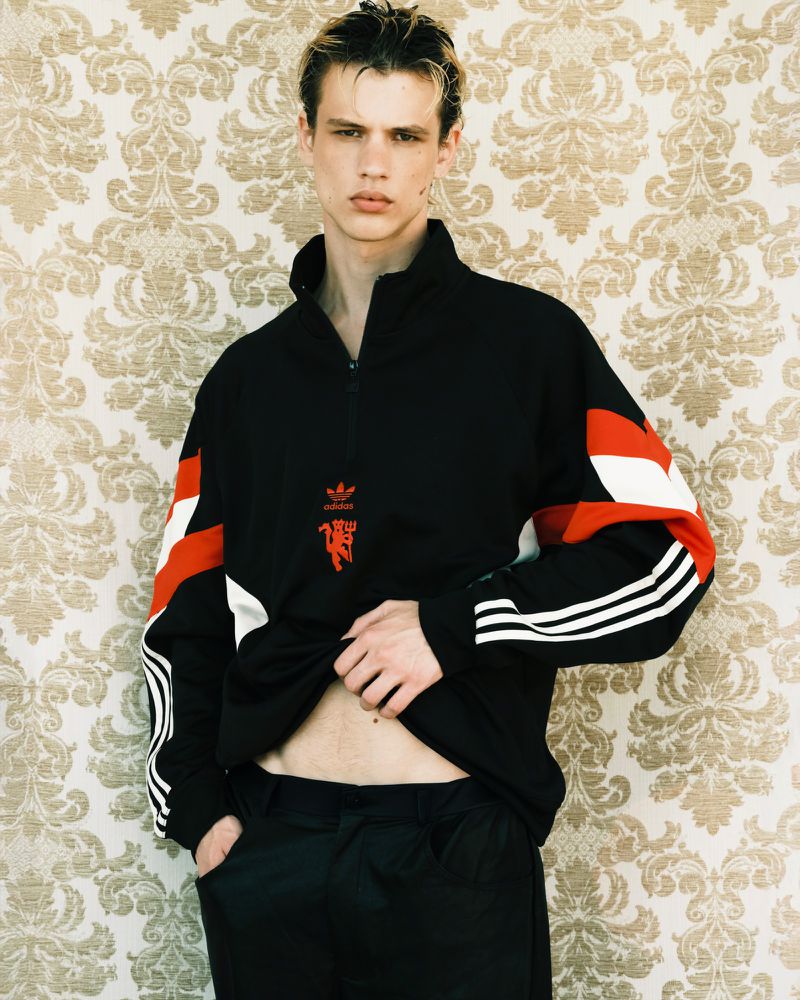 Model Lazar Dedic wearing a black adidas track jacket with red and white accents against ornate damask wallpaper.