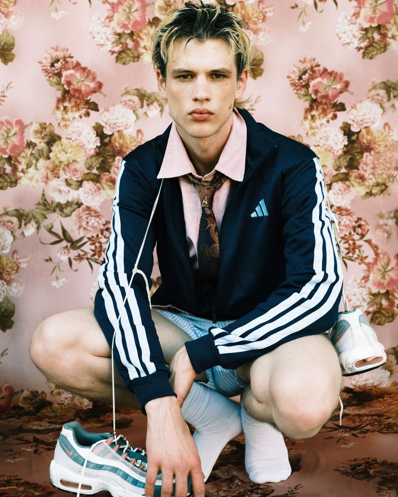 Man in athletic jacket and shorts posing with sneakers against a floral backdrop, wearing high socks and a patterned tie.