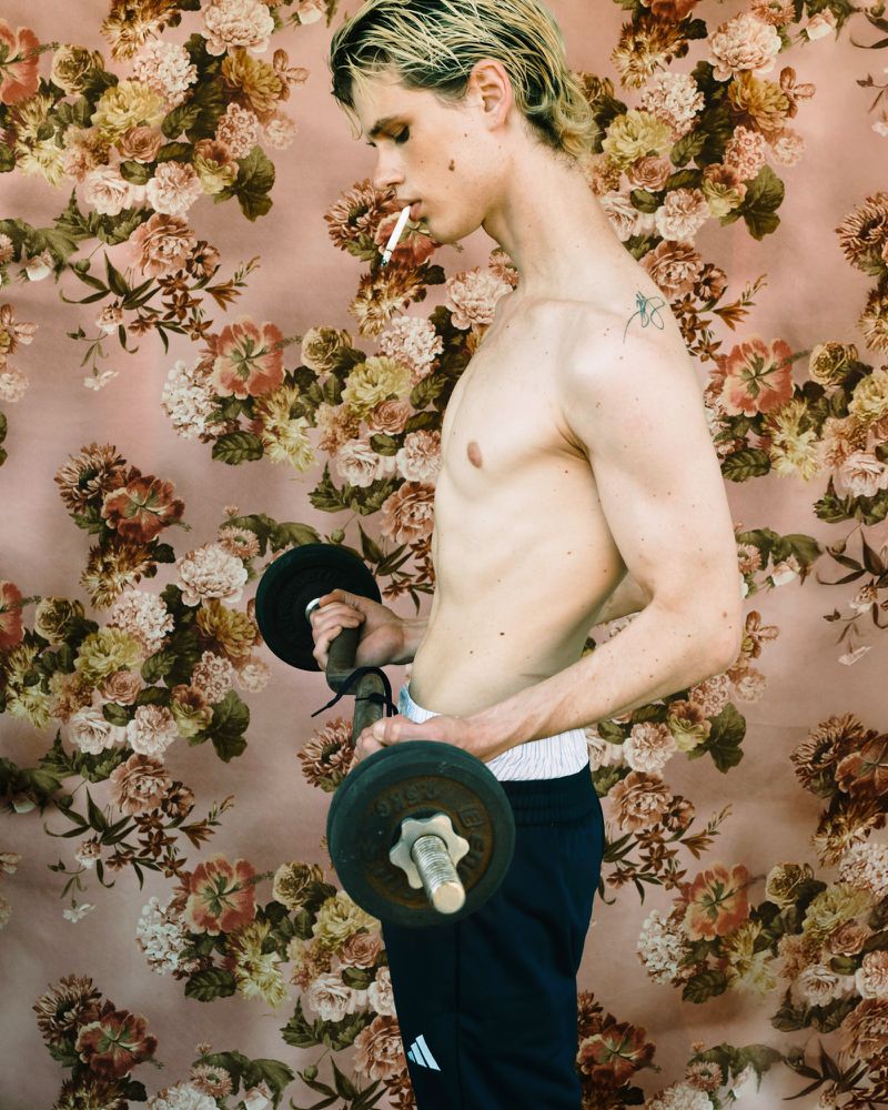 Shirtless man with platinum blonde hair lifts barbell, cigarette in mouth, set against floral wallpaper backdrop in a fitness themed portrait.