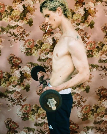 Shirtless man with platinum blonde hair lifts barbell, cigarette in mouth, set against floral wallpaper backdrop in a fitness themed portrait.