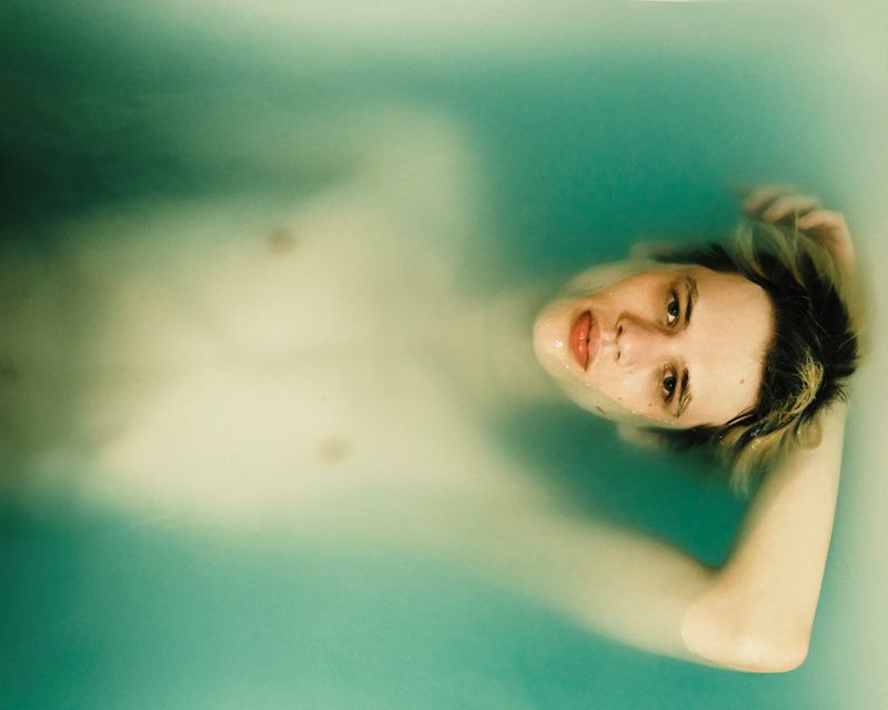 Person with wet blonde hair, partially submerged in a turquoise bathtub, gazes toward the camera in a close-up shot with diffused lighting.