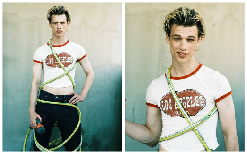 Young adult poses in white t-shirt, dark jeans, accessorized with a green garden hose wrapped around his body, outdoors against grey concrete.