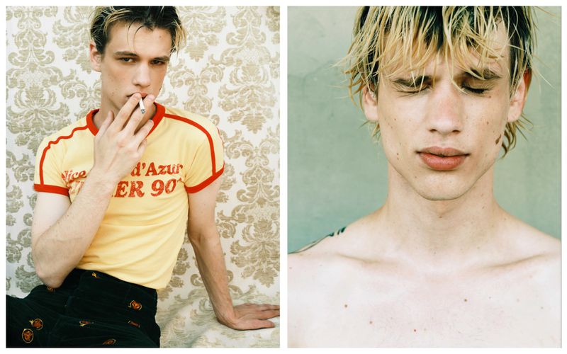Blonde male model in a yellow t-shirt with red trim smoking a cigarette against a floral wallpaper and light green backdrop.