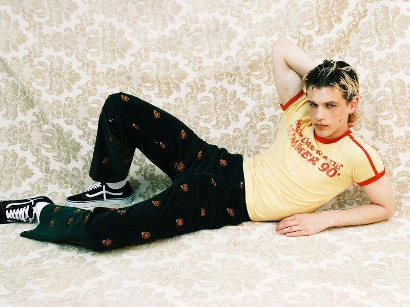 Young adult male with blond hair reclines on a floral patterned couch wearing a yellow t-shirt, dark pants, and sneakers in an indoor setting.