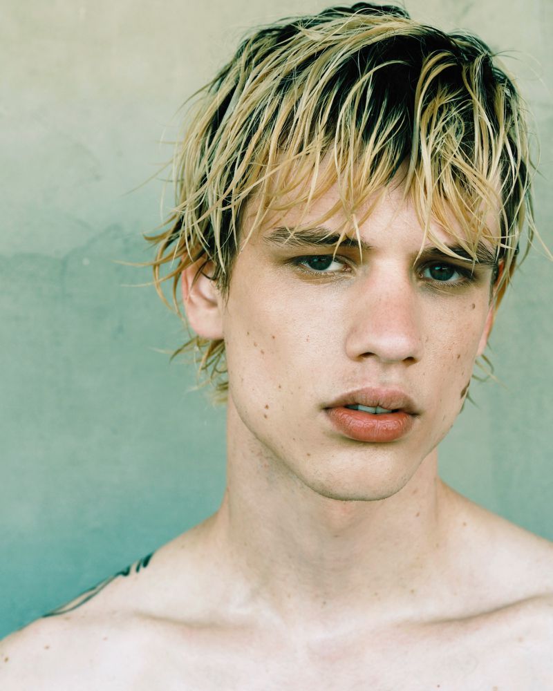 Close-up studio portrait of a young adult man with fair skin and blond hair, looking at the camera against a neutral background.