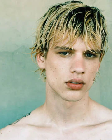 Close-up studio portrait of a young adult man with fair skin and blond hair, looking at the camera against a neutral background.
