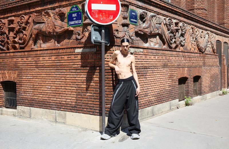 Young adult poses in baggy pants on a sunny Paris street corner next to a red and white Do Not Enter sign and building with carvings.