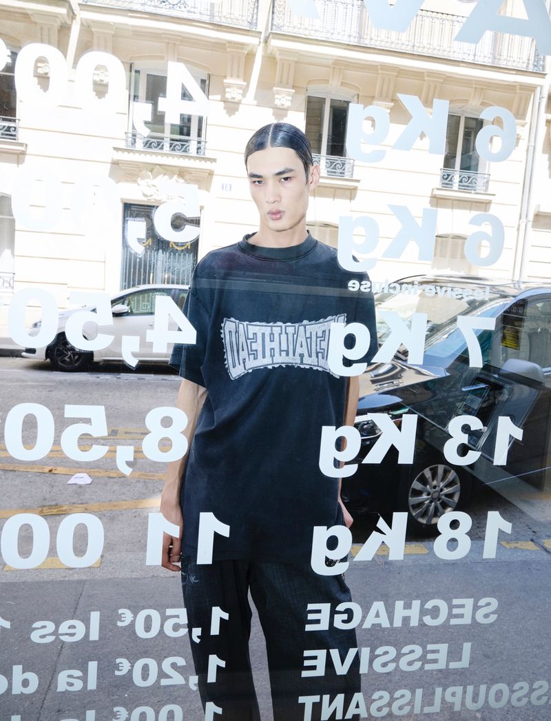 Adult male poses wearing black t-shirt and pants. Building and car are visible through window with signage in an urban setting.