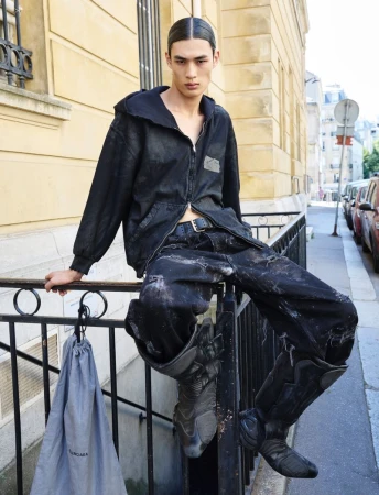 An Asian male model poses on a Parisian street wearing a black jacket, distressed jeans, and tall leather boots with a Balenciaga bag.