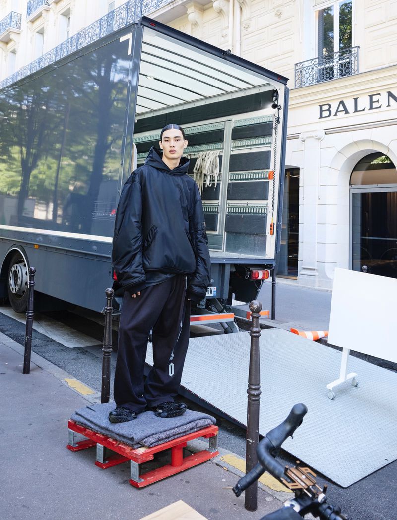 Fashion shoot of a person wearing a black hoodie and baggy pants standing on a platform in front of a truck on a city street.