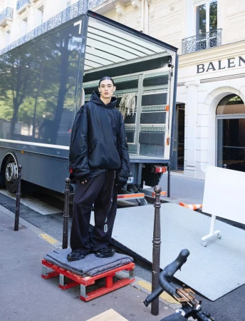 Fashion shoot of a person wearing a black hoodie and baggy pants standing on a platform in front of a truck on a city street.