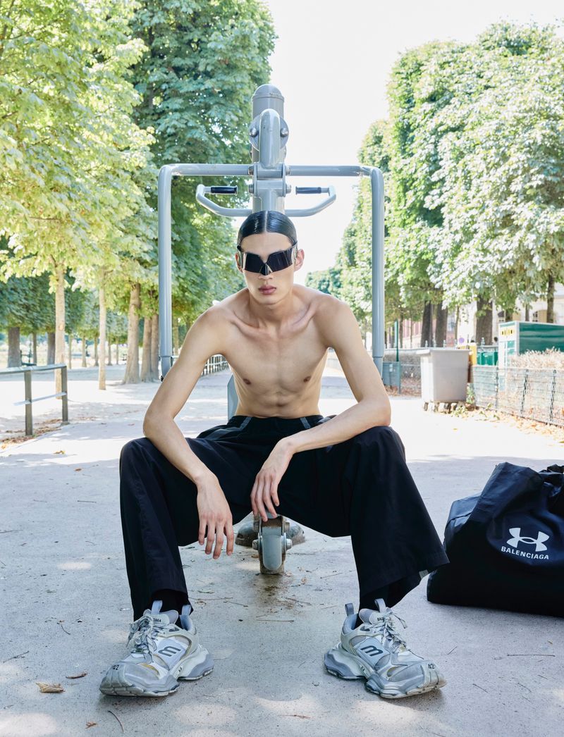 An adult male poses shirtless on exercise equipment in a park, wearing sunglasses and black pants, with a duffle bag nearby.
