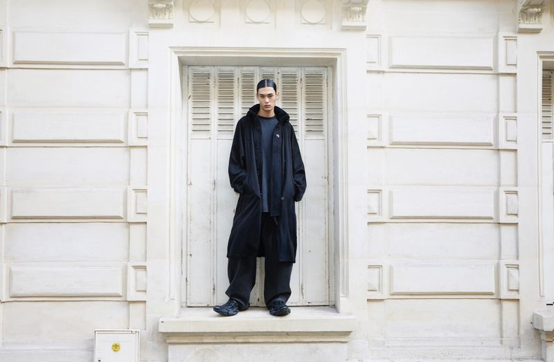 Fashionable young adult in long black coat and pants posing on a doorstep in front of a white shuttered door against a white building.