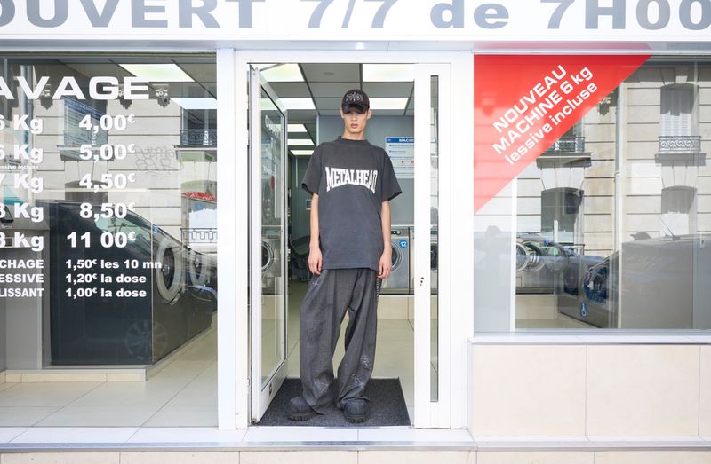 An adult male poses in front of a laundromat, wearing an oversized gray t-shirt and sweatpants. The laundromat has washing machines inside.