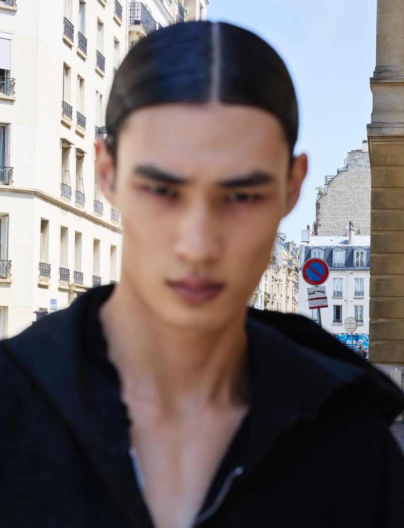 Close-up of an Asian male model with slicked-back hair in a black hoodie posing in an urban setting with buildings and a blue sky.