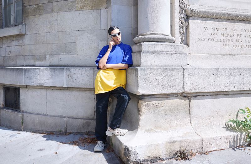 Adult female posing near a stone building wearing a blue t-shirt, yellow apron, black pants, white sneakers, and white sunglasses.
