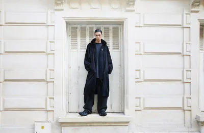 Model in black jacket and dark trousers standing in a recessed doorway against an off-white stone building facade.