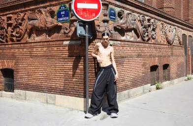 Model Zhaoyi Fan standing shirtless in wide-leg black trousers next to a street sign against an ornate brick building in Paris.