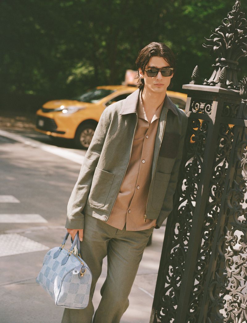 Man in an outdoor setting wearing sunglasses, a jacket and pants, carrying a patterned bag with a yellow taxi in the background.