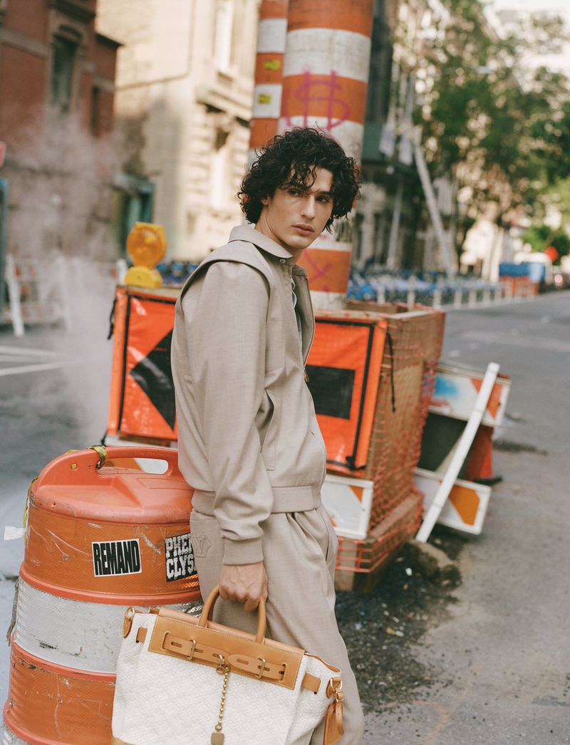 Man in a beige tracksuit with a patterned satchel stands on a city street with construction barrels in the background on a sunny day.