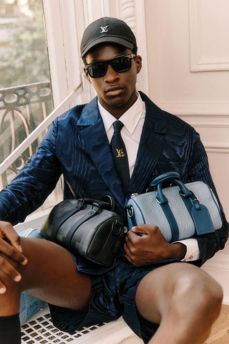 A stylish Black man sits indoors with designer bags, wearing a blue suit, sunglasses, and a baseball cap. He models a luxury fashion look.
