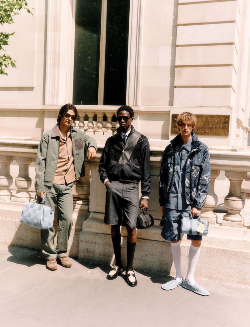 Three adult male fashion models pose outdoors in jackets, shorts, and loafers, holding bags, against a light stone wall backdrop.