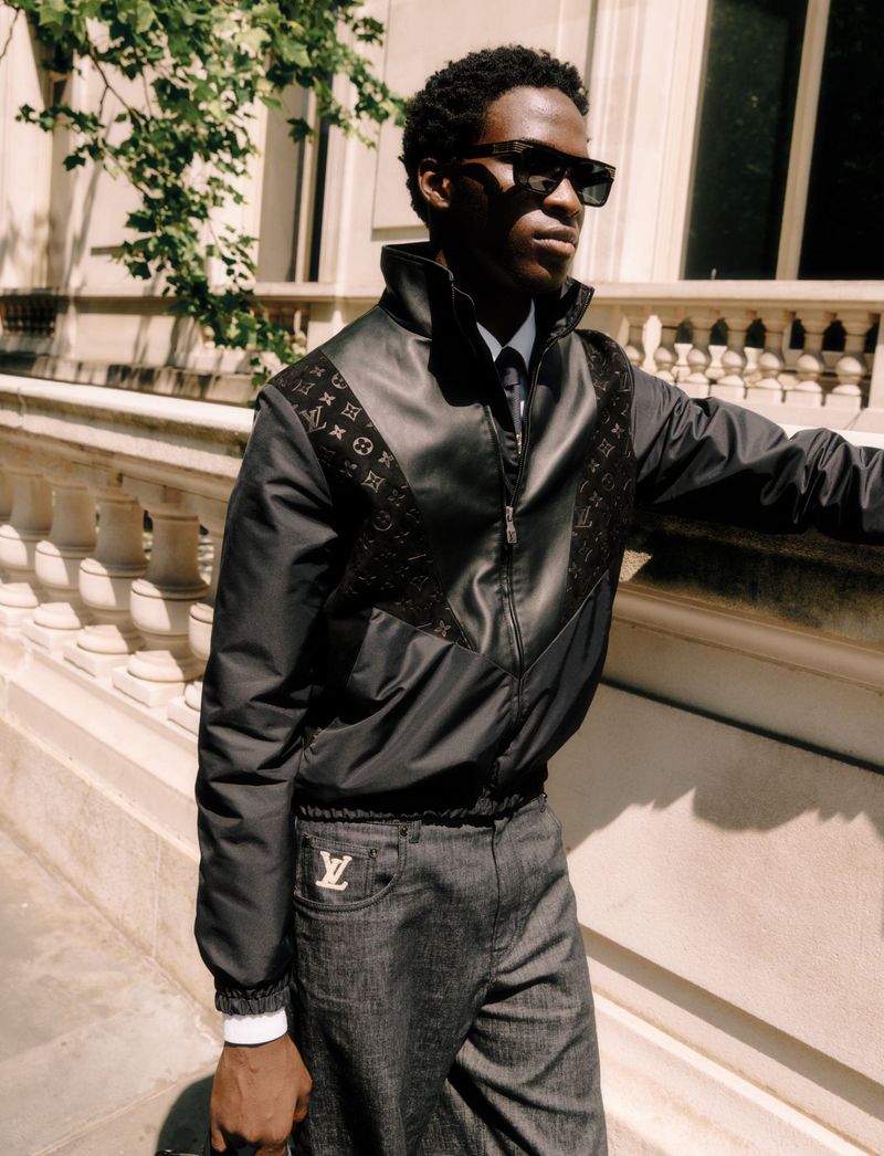 A dark-skinned male fashion model in a jacket with monogram details poses outdoors against a stone wall with trees and natural light.