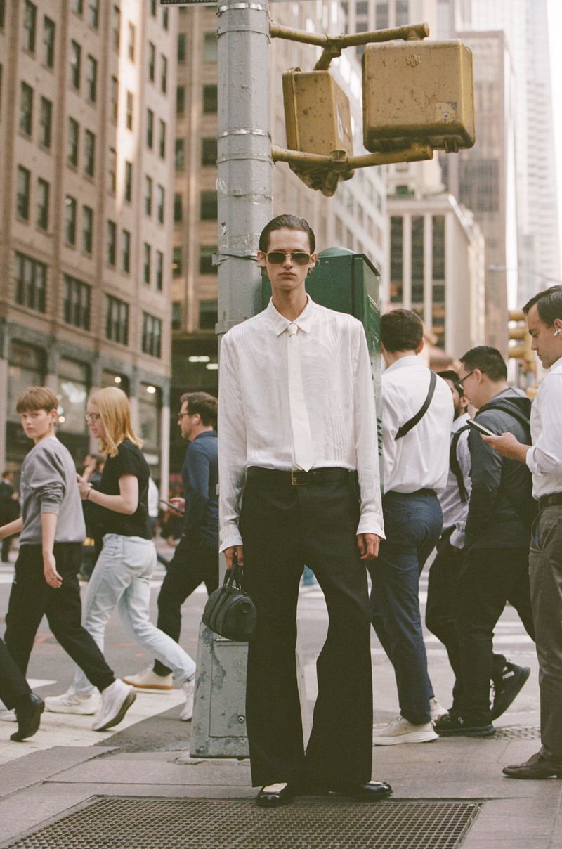 Fashionable man poses on a city sidewalk next to a light pole with a leather bag, wearing sunglasses, a button-down shirt, and black trousers.