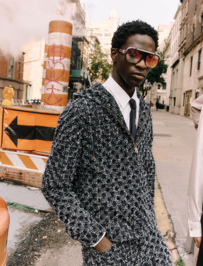 A young Black man poses on a city street in a stylish tweed jacket, white shirt, and tie, sporting aviator sunglasses with smoke rising in the background.