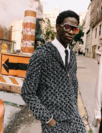 A young Black man poses on a city street in a stylish tweed jacket, white shirt, and tie, sporting aviator sunglasses with smoke rising in the background.