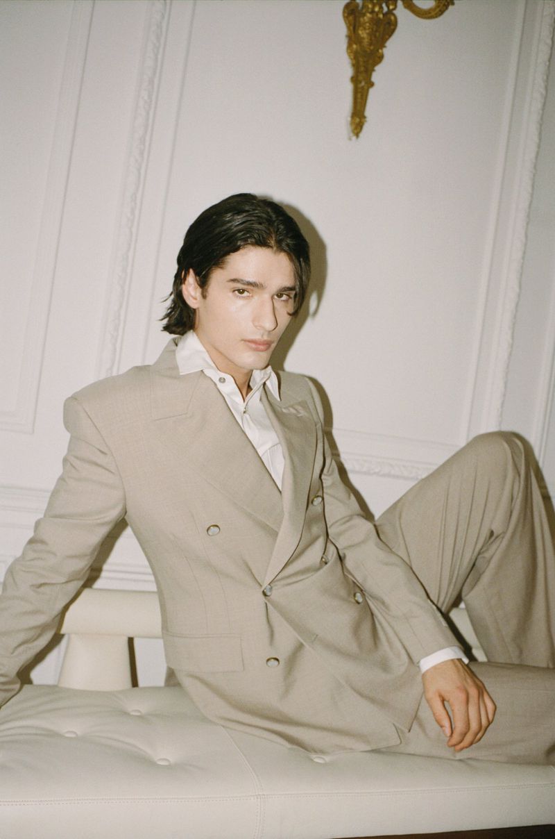 Young adult man in a beige suit and white shirt poses on a tufted white bench in a bright indoor setting with gold wall accents.
