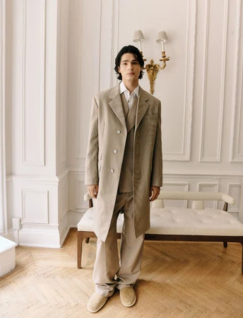 A young adult male posing in a beige suit and coat in a light-filled interior room, creating a timeless fashion portrait.