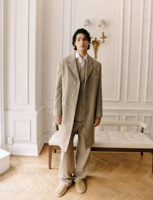 Male model in an oversized beige overcoat and matching trousers standing in an interior room with herringbone wood floors.