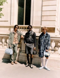 Three male models posing outdoors in front of a classical building exterior holding designer handbags