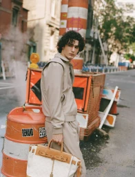 Model posing in a beige matching jacket set while holding a structured tan and cream tote bag next to orange traffic barrels.