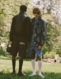 Two male models standing outdoors on a grassy park lawn, wearing designer menswear with monogram jackets and shorts.