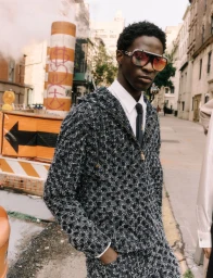 Black male model wearing oversized tinted sunglasses, a textured tweed hooded jacket, and a tie on an urban street.