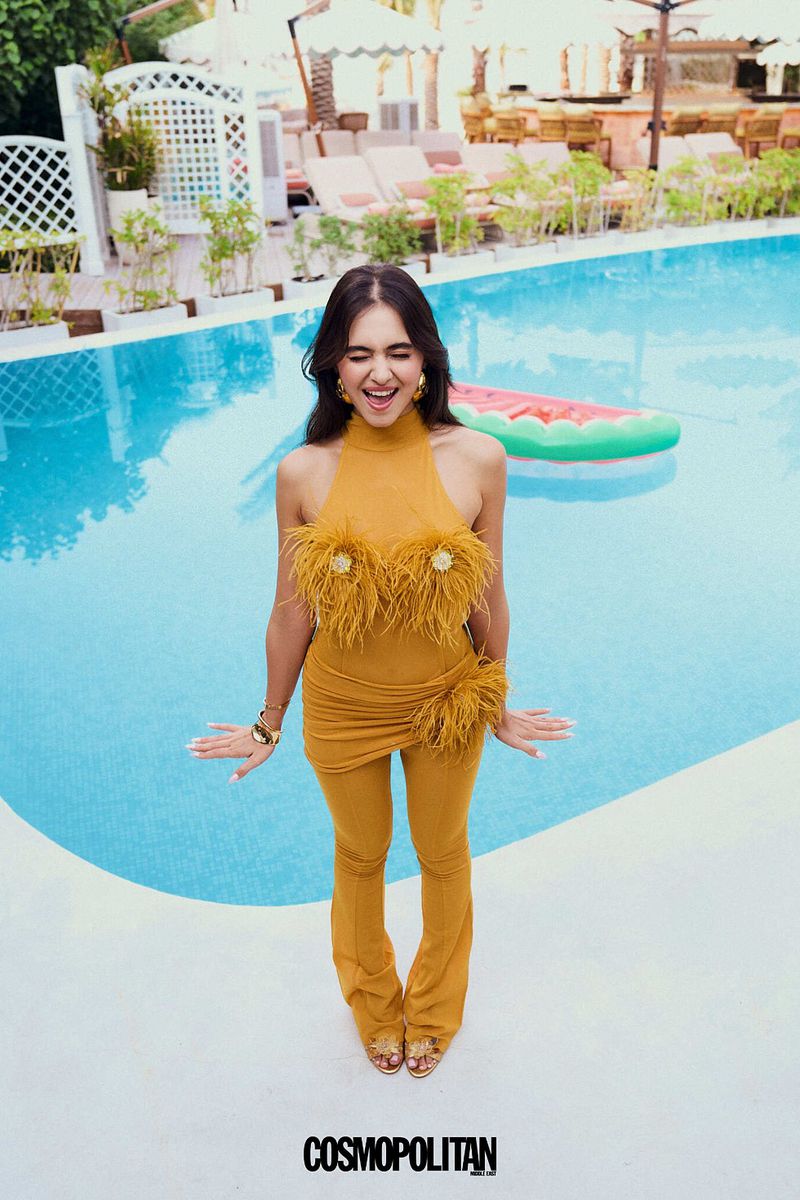 Young woman in mustard-colored feather-trimmed outfit smiling and posing outdoors by a pool and lounge area on a sunny day.