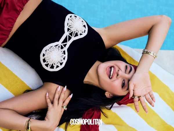 Smiling woman in black top with white embellishments, lying on striped towel near pool, accessorized with bracelets and rings, warm tones.