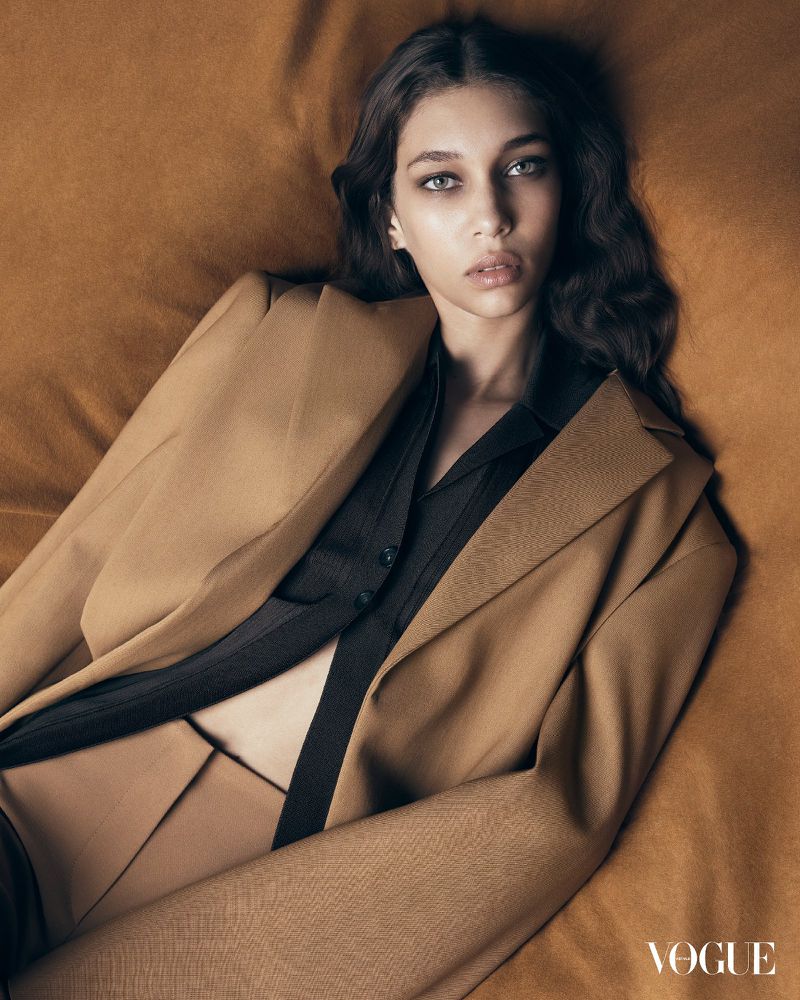 Woman wearing a blazer and button-down shirt lies on a bed in a studio portrait for Vogue magazine. Her gaze is directed at the camera.