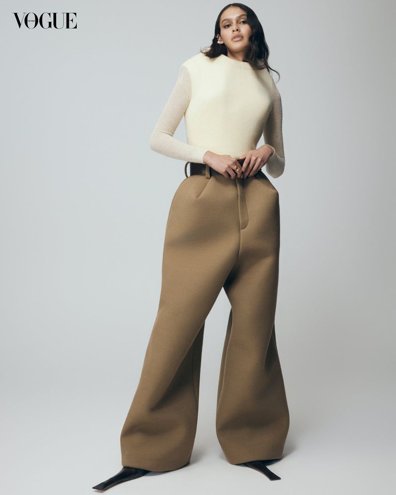 Fashion editorial shot: A woman in a cream top and bulbous brown trousers poses in a photo studio against a neutral background. Vogue logo visible.