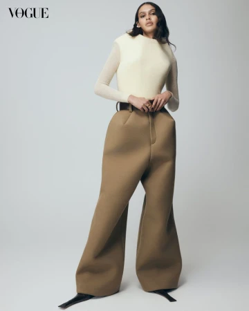 Fashion editorial shot: A woman in a cream top and bulbous brown trousers poses in a photo studio against a neutral background. Vogue logo visible.