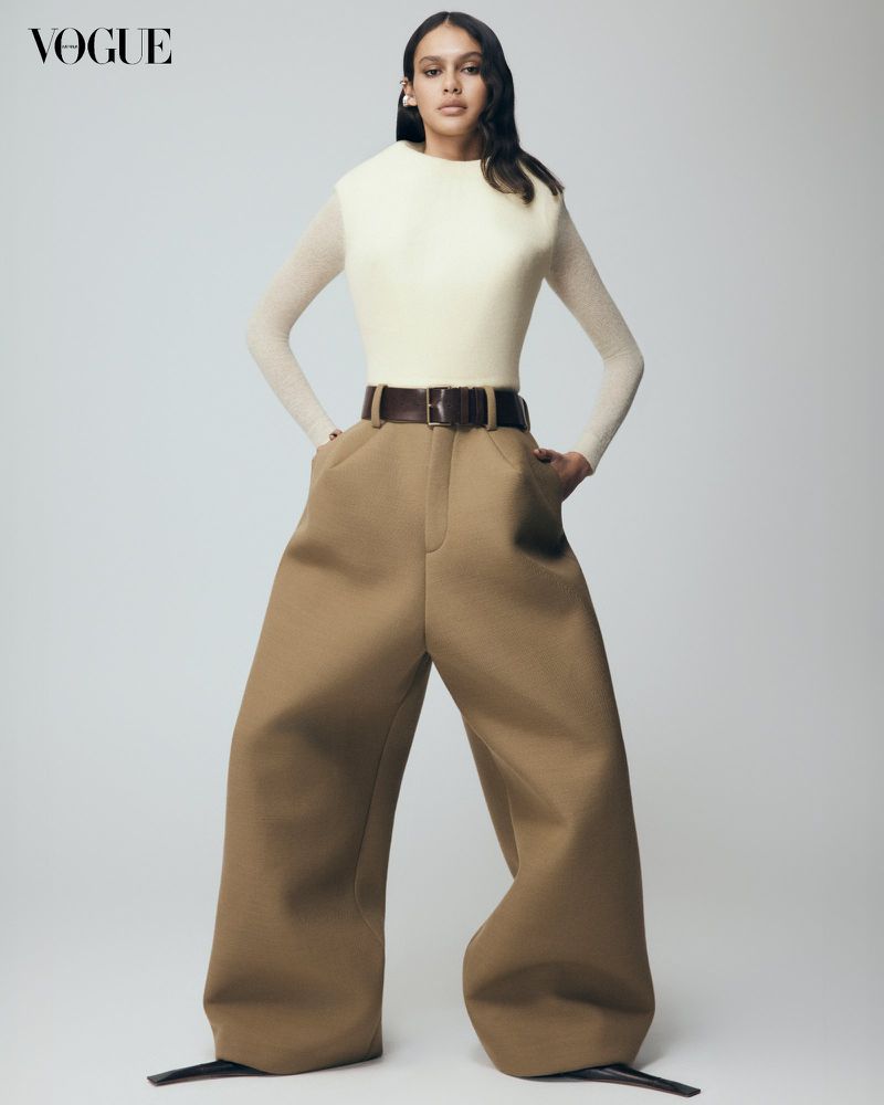 Full-length fashion portrait of a woman posing in oversized pants, a cream top, and a dark brown belt in a photo studio.
