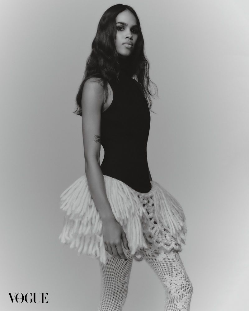 A woman in a black top, white tutu-style skirt, and lace leggings poses in a studio with a Vogue logo on a neutral background.