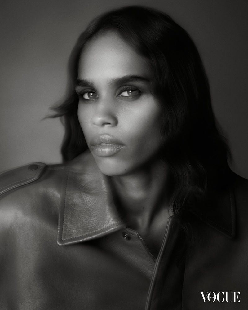 Portrait of an adult female of mixed ethnicity with long dark hair, wearing a leather jacket, in a black and white indoor studio shot.