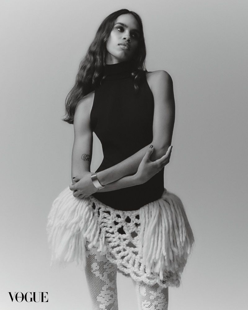 Black and white portrait of a woman in a black top, a white skirt with fringes, and patterned white tights standing in a studio.