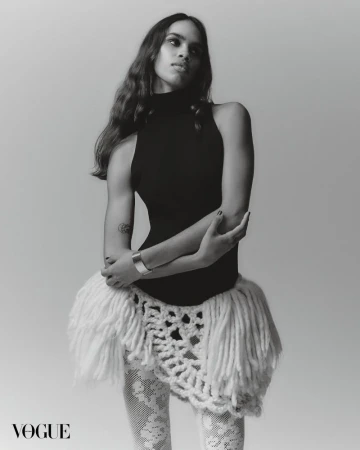 Black and white portrait of a woman in a black top, a white skirt with fringes, and patterned white tights standing in a studio.