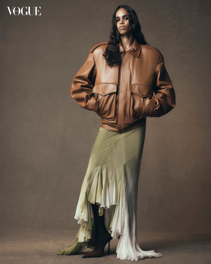 A model poses in a tan leather bomber jacket and long flowing skirt against a neutral background for a fashion editorial.