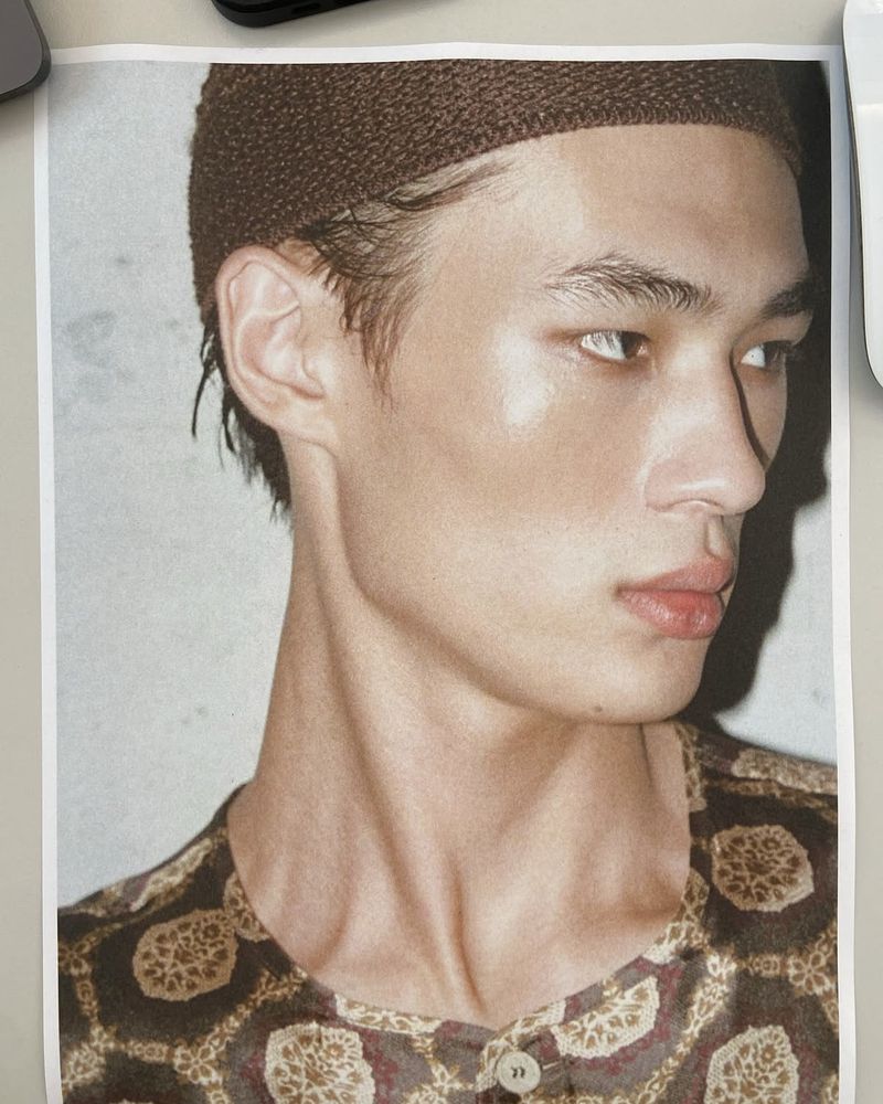 Portrait of a young Asian man wearing a brown cap and patterned shirt, looking off to the side, photographed indoors with natural lighting.